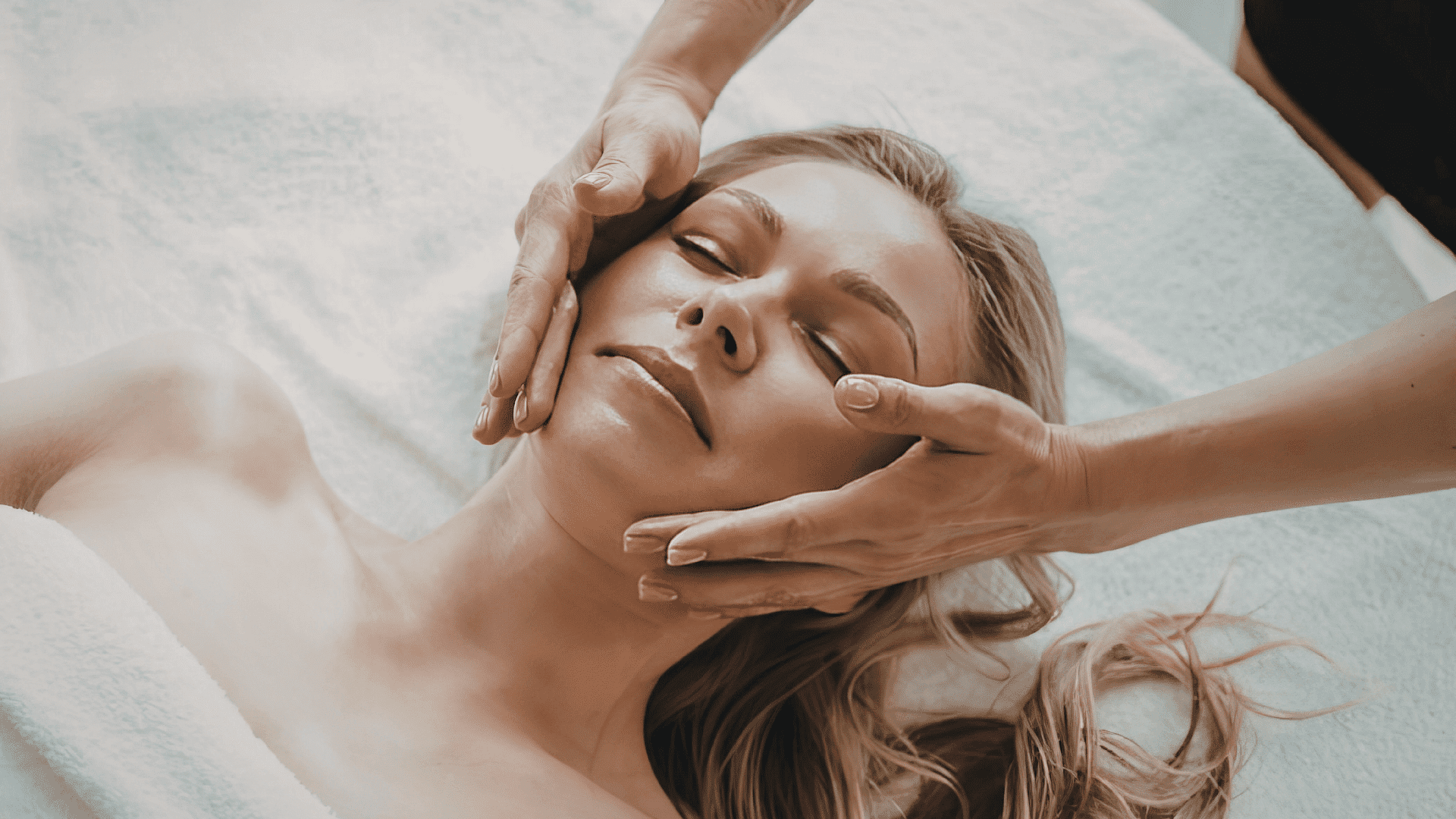 Facial massage at a spa, woman relaxed on a soft towel with eyes closed.