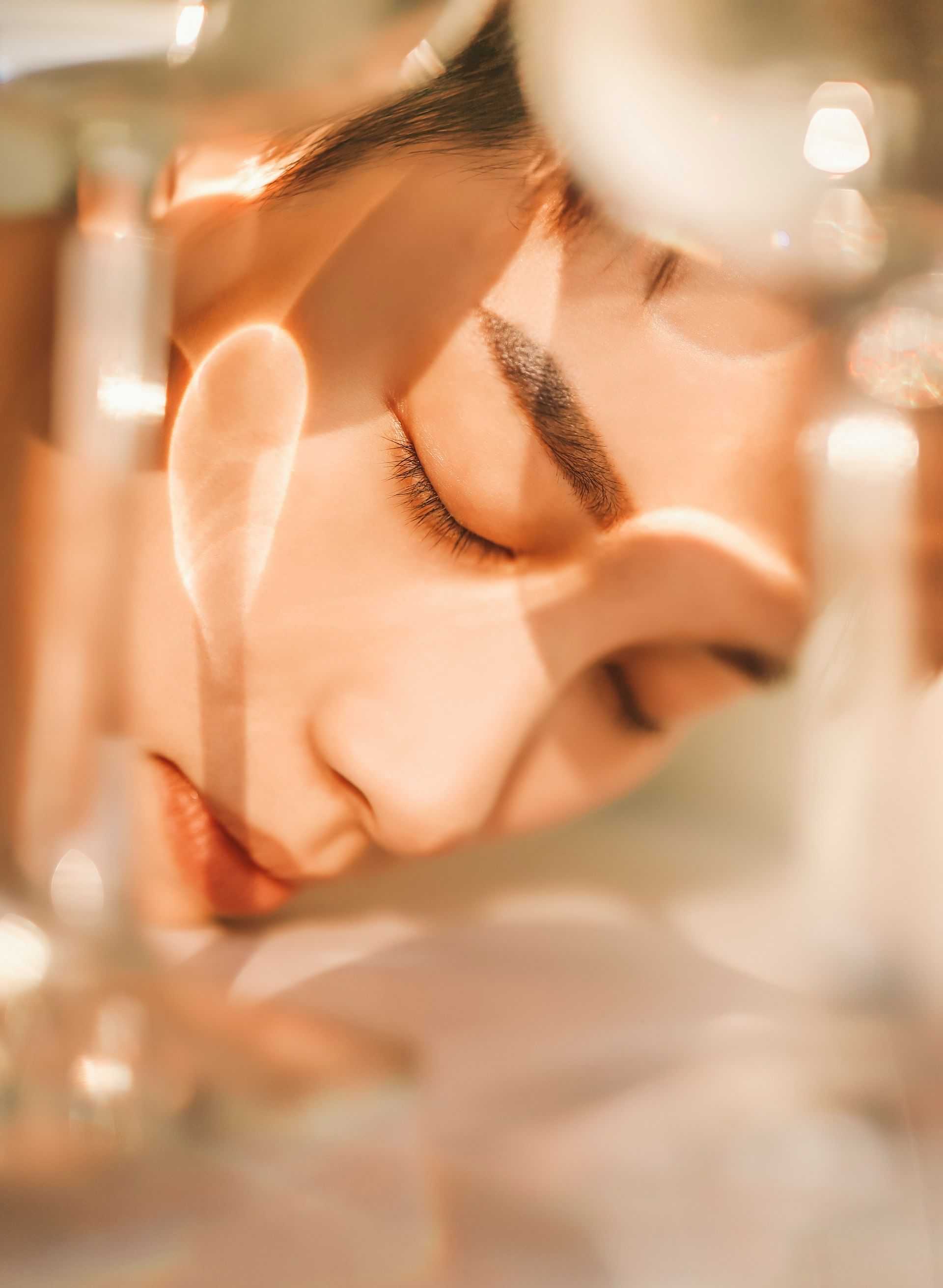 Close-up of a serene person with eyes closed, surrounded by soft, warm lighting and glass reflections.