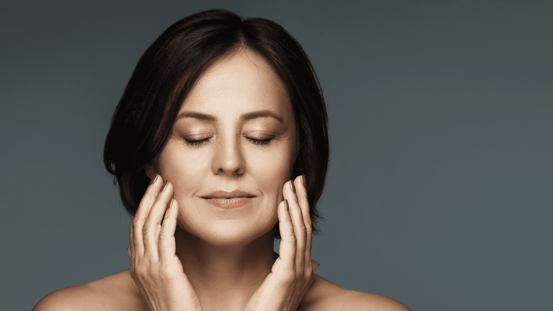 Woman with serene expression, eyes closed, hands gently touching her face against gray background.